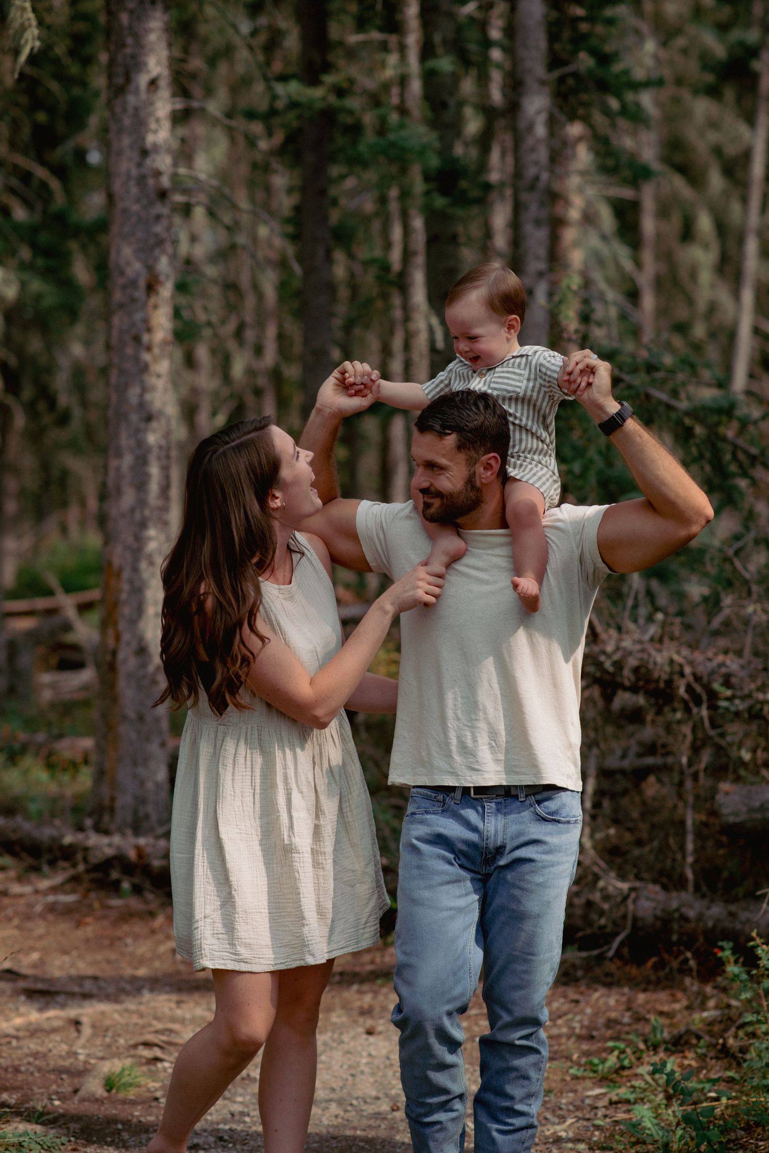 Professional family photos Alberta, father carrying baby boy on shoulders as mom engages with him in forest