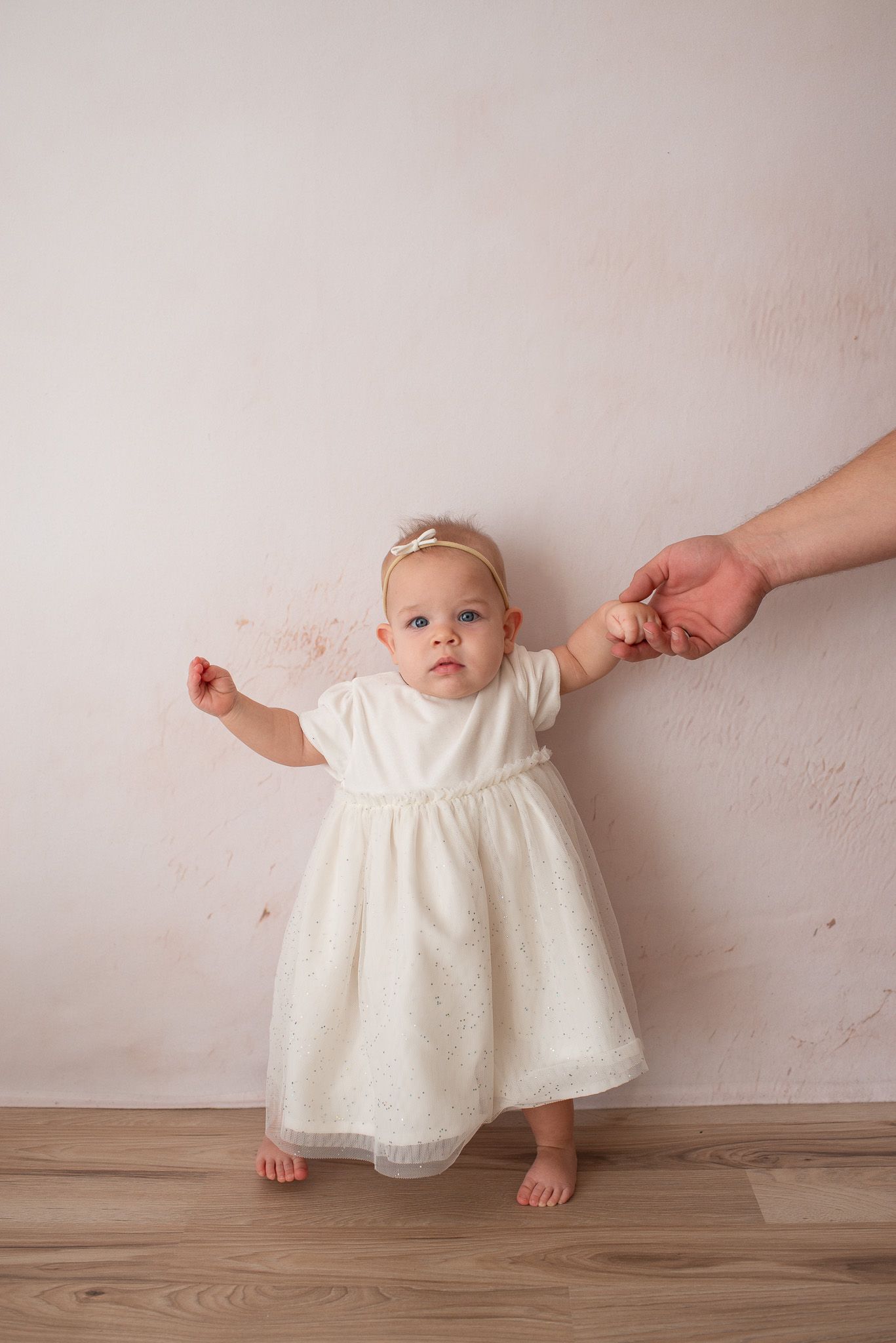 Baby wearing white lace dress sitting on wooden floor while hands support from both sides.