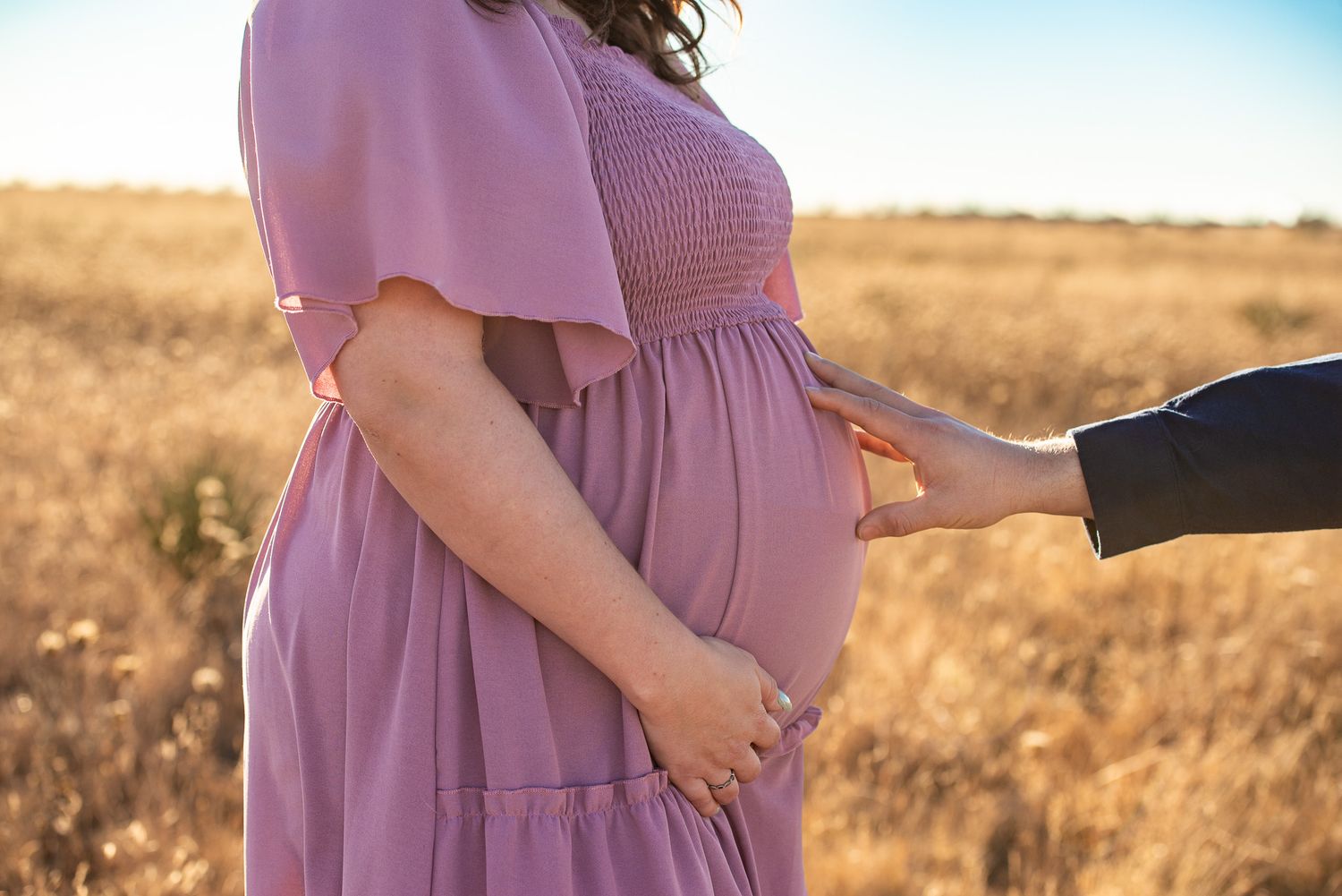 A maternity photoshoot in a golden field at sunset with woman in flowing mauve dress.