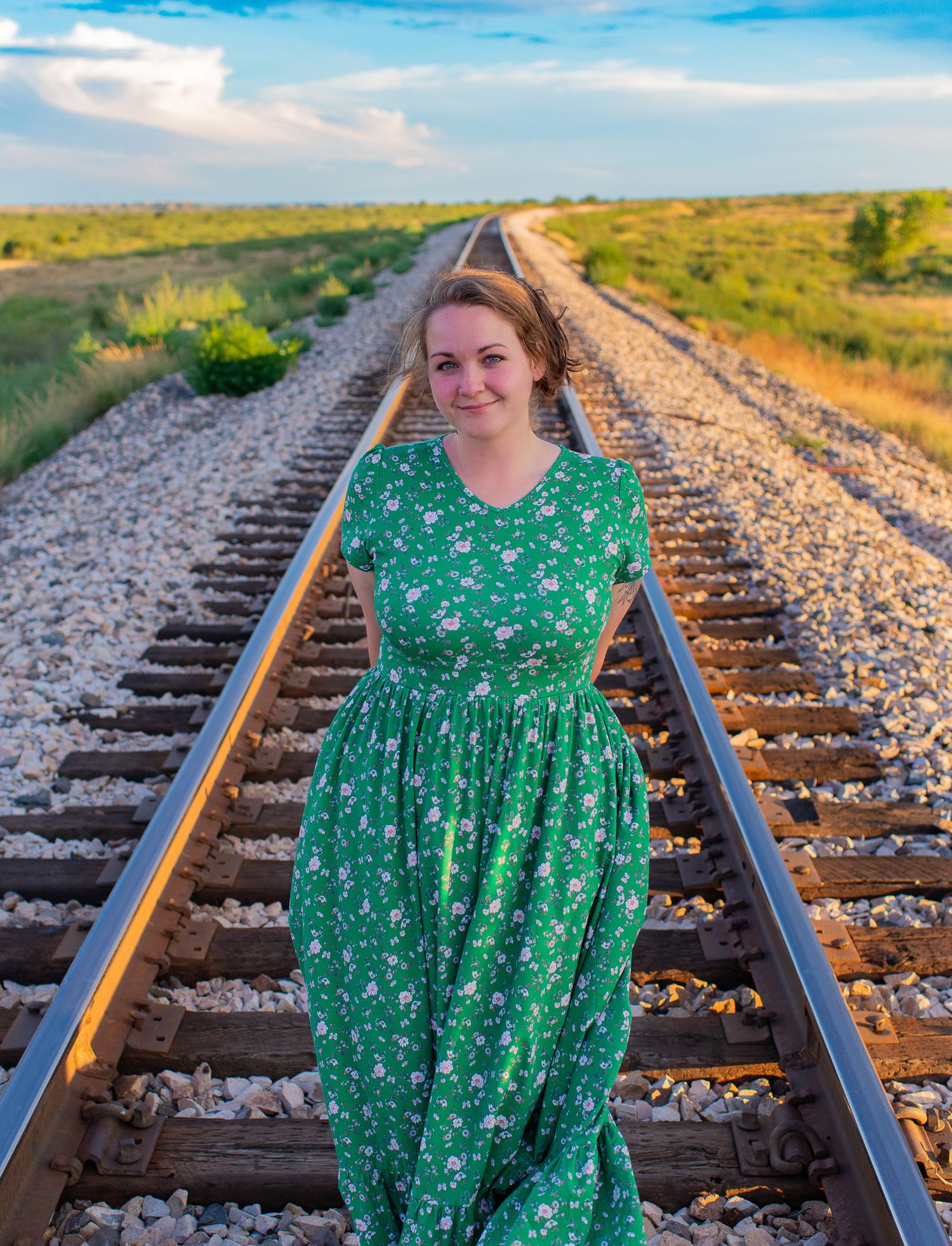 Person in green floral dress poses along railroad tracks at sunset.