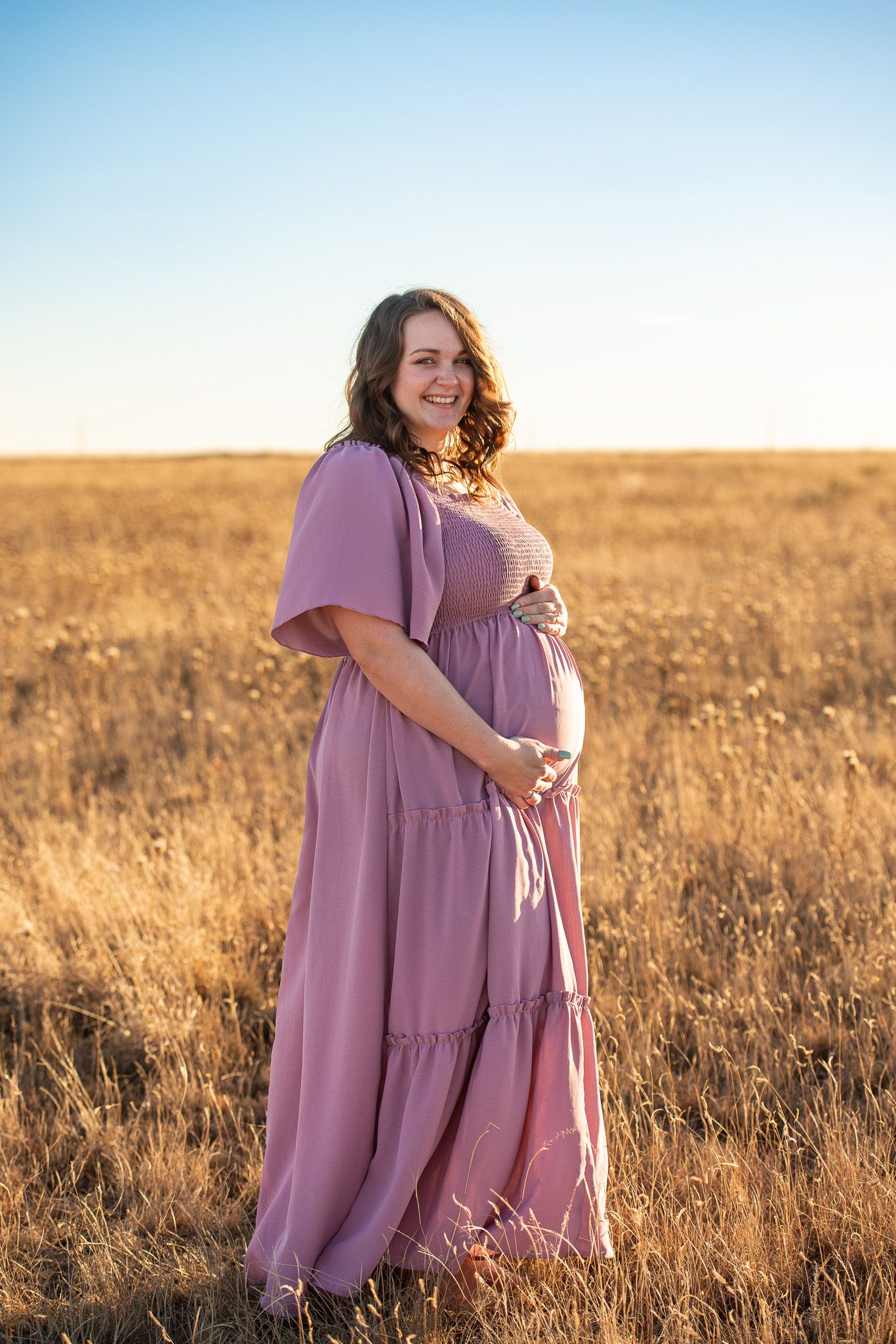 Woman in flowing mauve dress poses in golden field during sunset photoshoot.