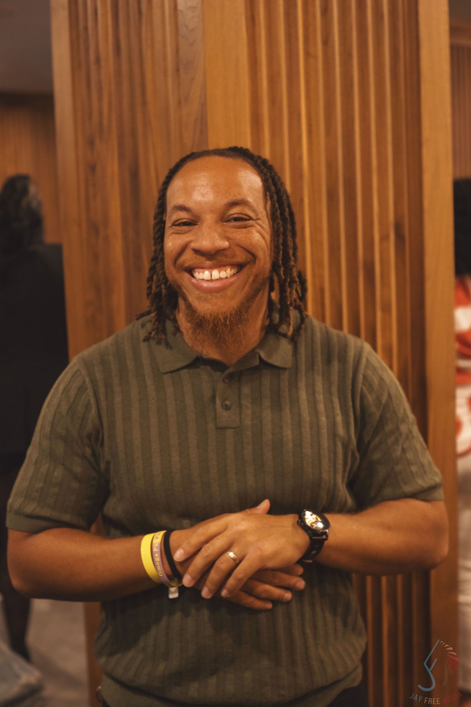 A person smiles warmly while wearing a dark striped shirt at a wooden paneled table.
