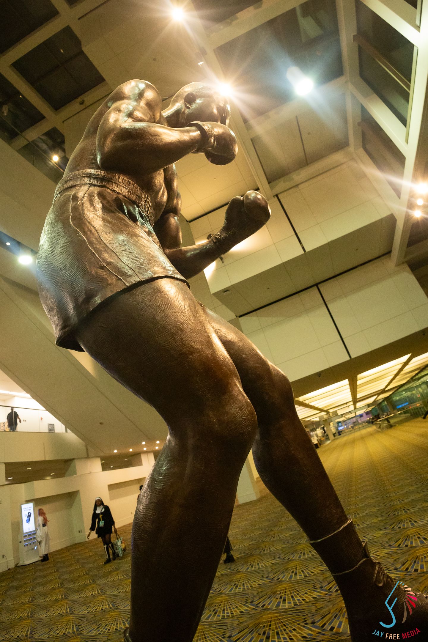 Bronze statue of a boxer in fighting stance displayed inside a large convention center with bright overhead lights.
