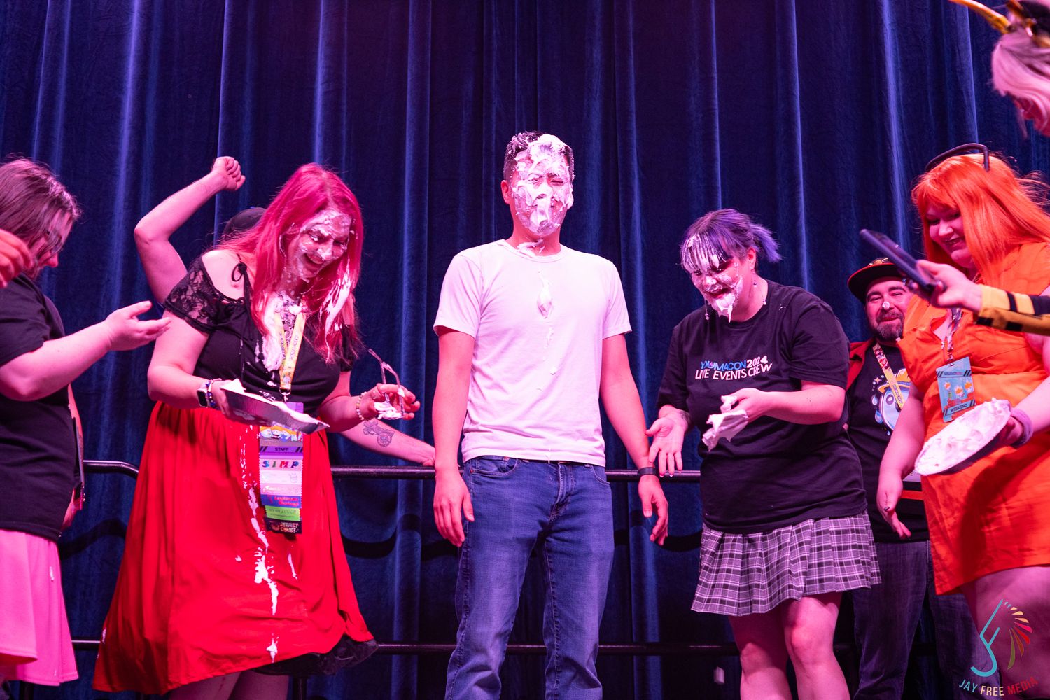 Group of cosplayers on stage at an anime convention event with pie on one participant's face.