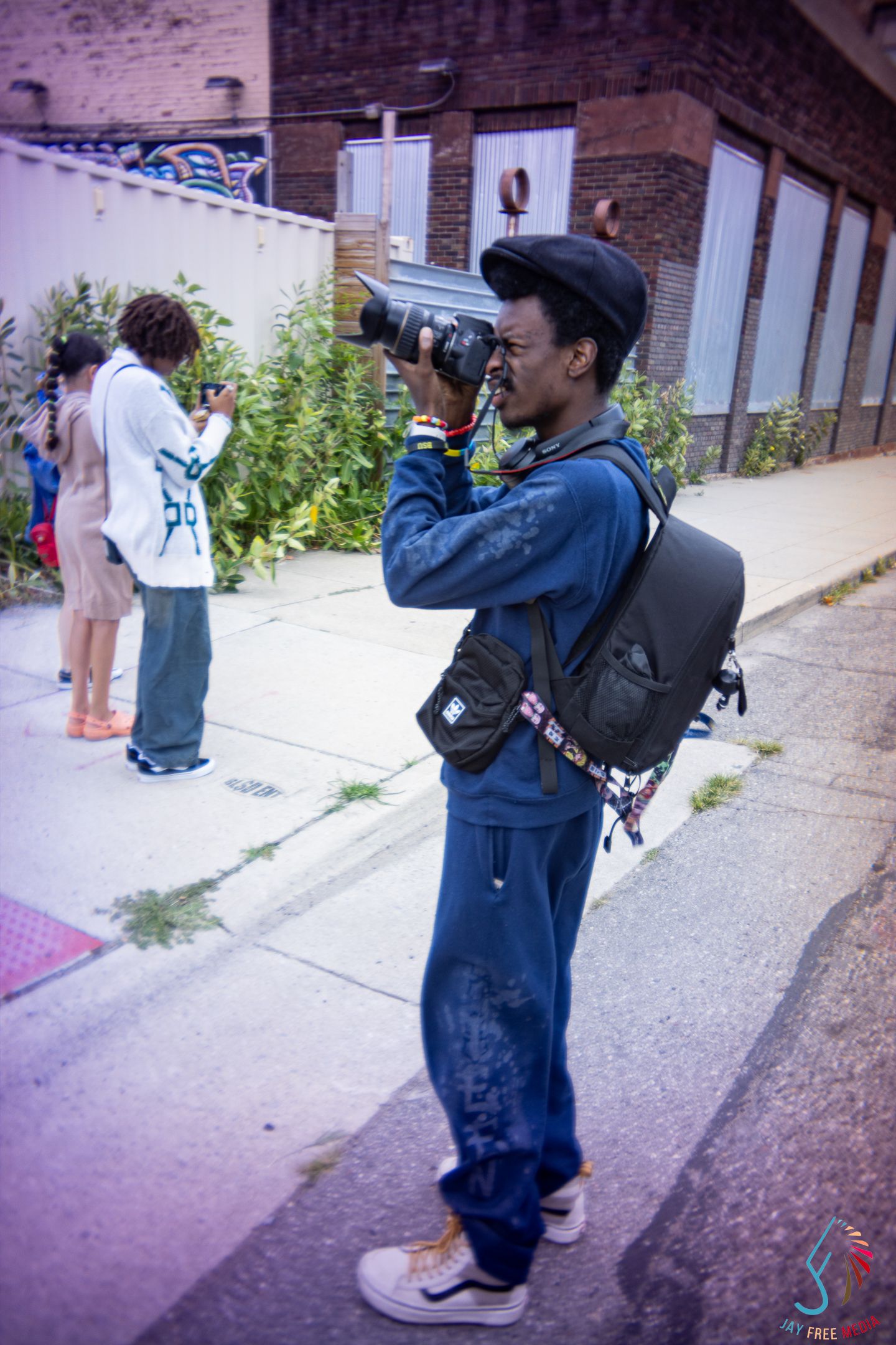 Young Black photographer with backpack shoots with a large telephoto lens on a city sidewalk.