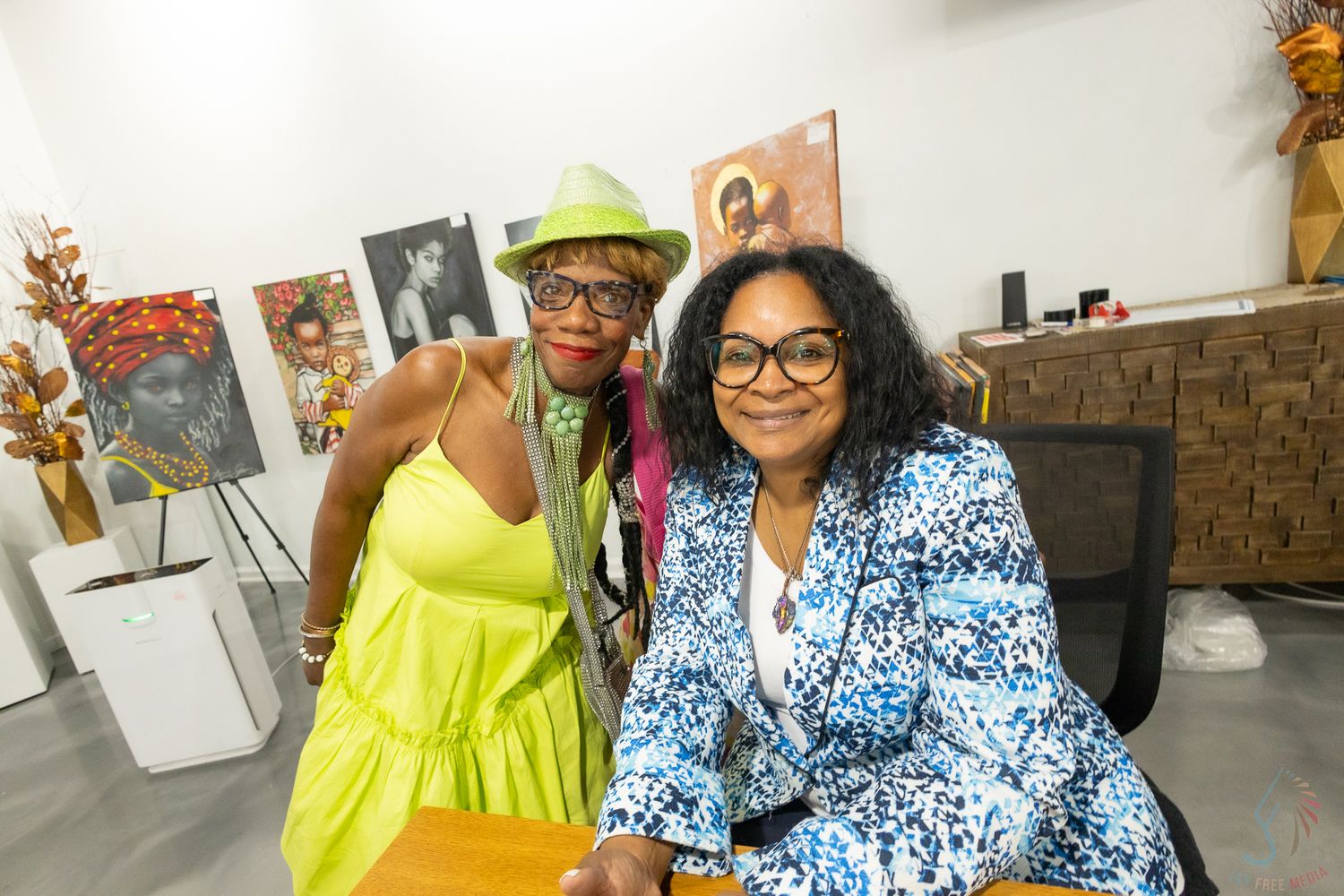 Two women smile together at an art gallery, one in neon yellow dress and green hat, other in blue floral blazer.