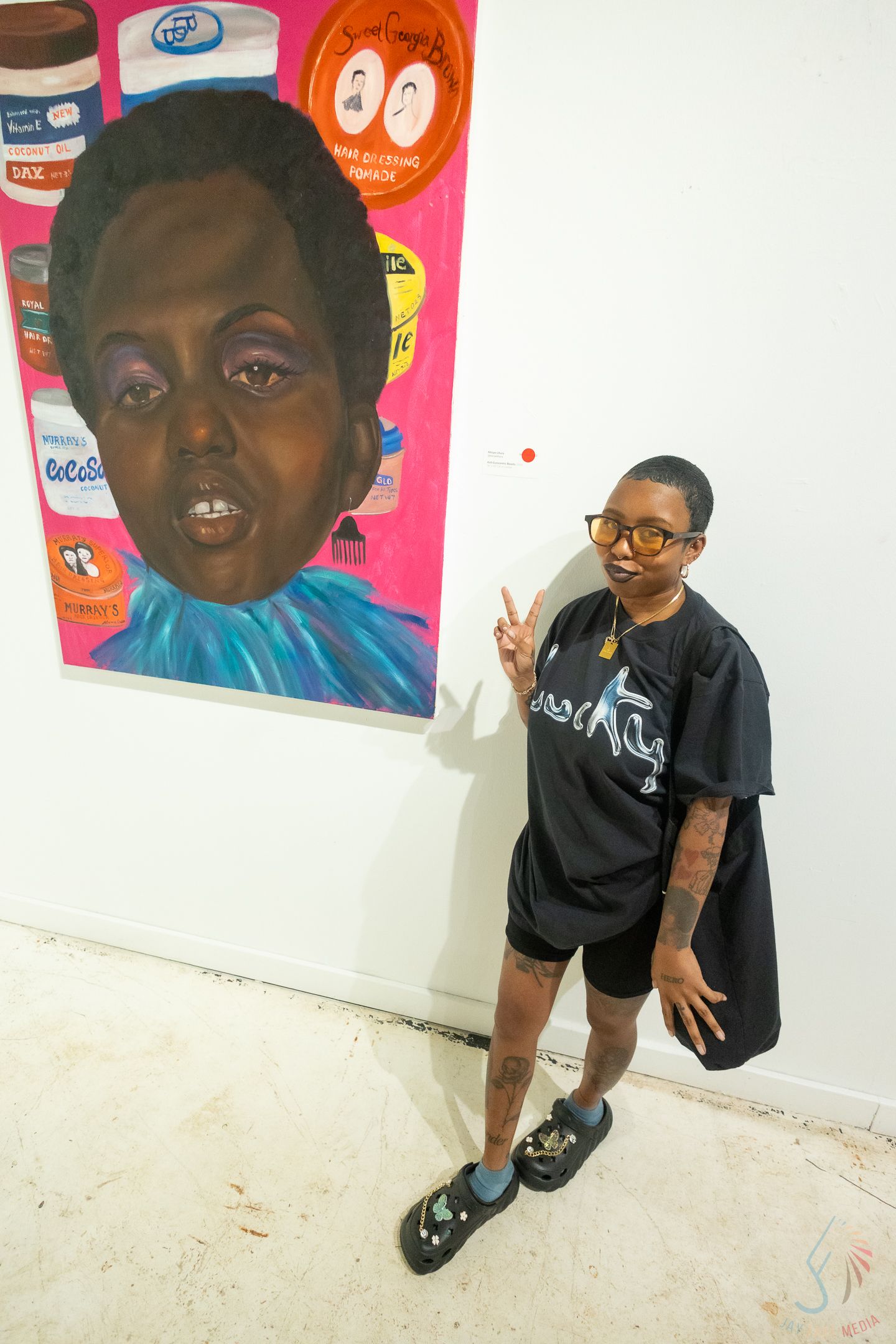 Artist poses with peace sign beside colorful portrait painting of a child surrounded by product labels on gallery wall.