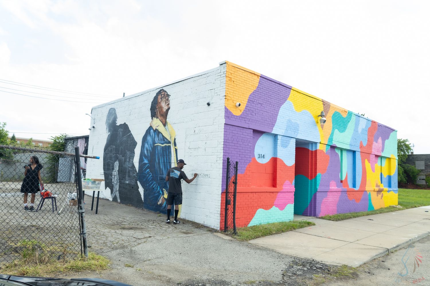 Colorful street mural on white brick building corner featuring figures and abstract shapes in vibrant hues.
