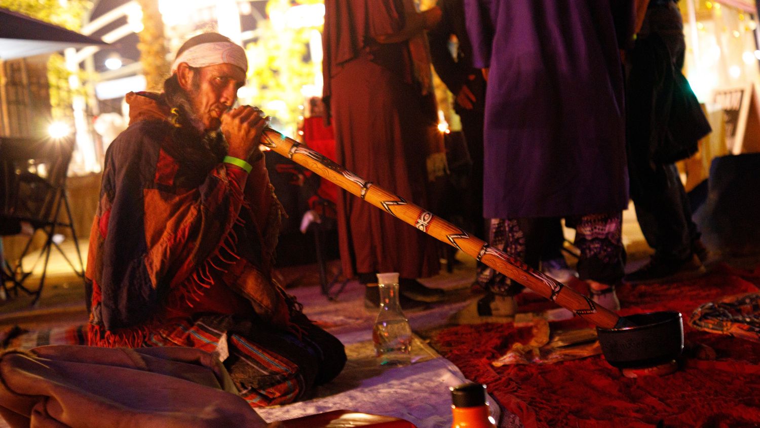 Musician playing a didgeridoo while seated on a colorful rug at a nighttime festival.