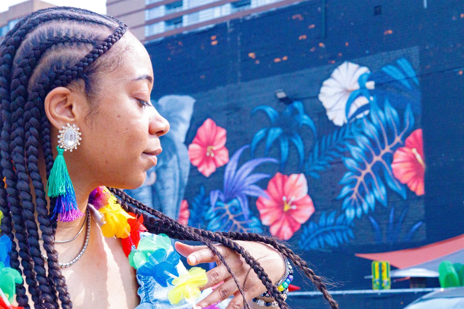 Young woman with colorful braids poses in front of a vibrant tropical floral mural wearing a rainbow lei.