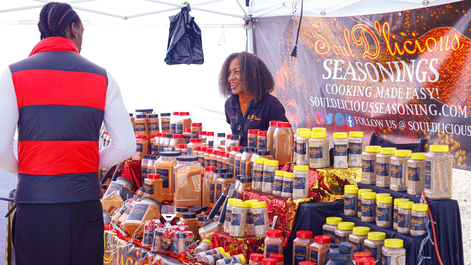 Vendor selling colorful spice jars at Sol Vicious Seasonings booth at outdoor food market.
