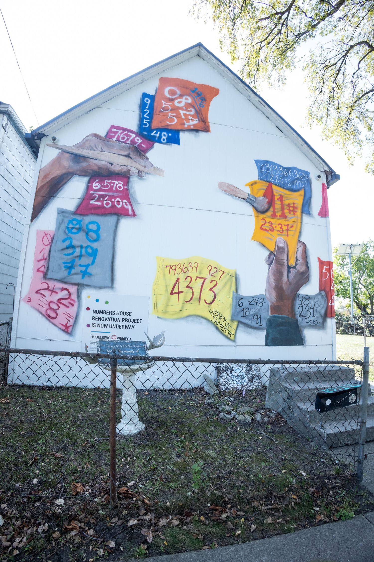 Colorful mural of hands holding numbered tags painted on the side of a white house during a renovation project.