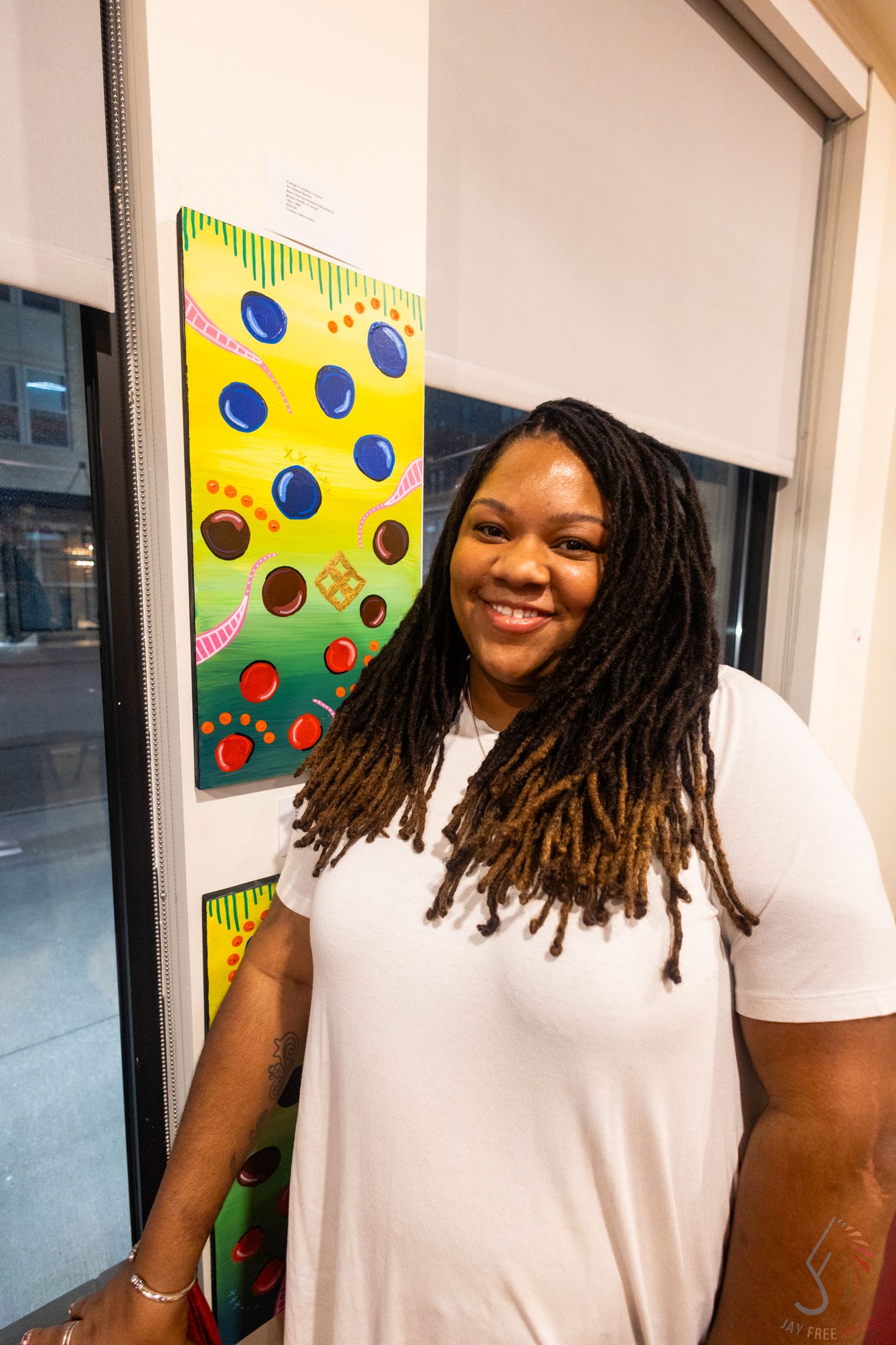 Smiling woman with long locs in white dress stands beside colorful abstract painting at art gallery.