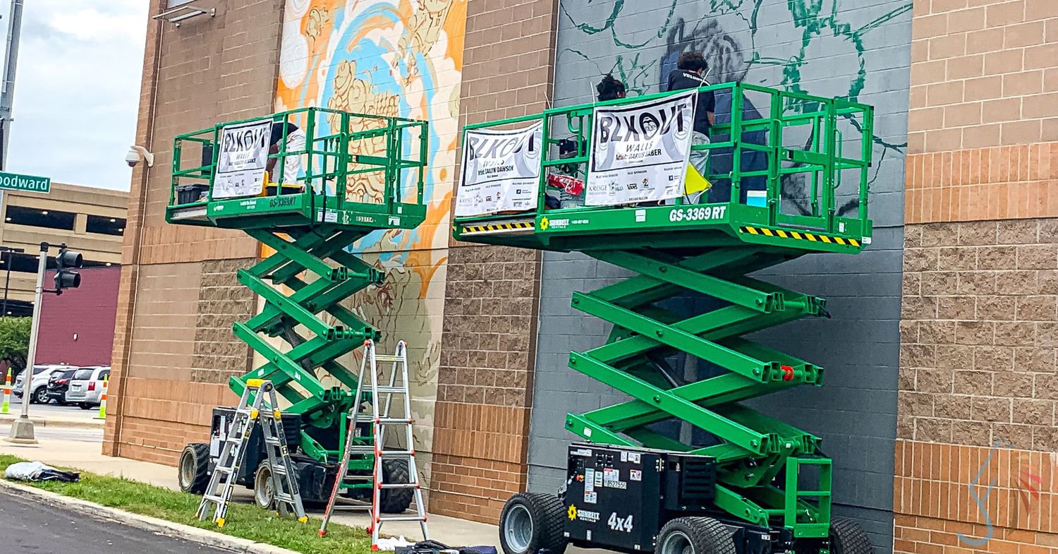 Workers on green scissor lifts painting a large colorful mural on a brick building exterior.