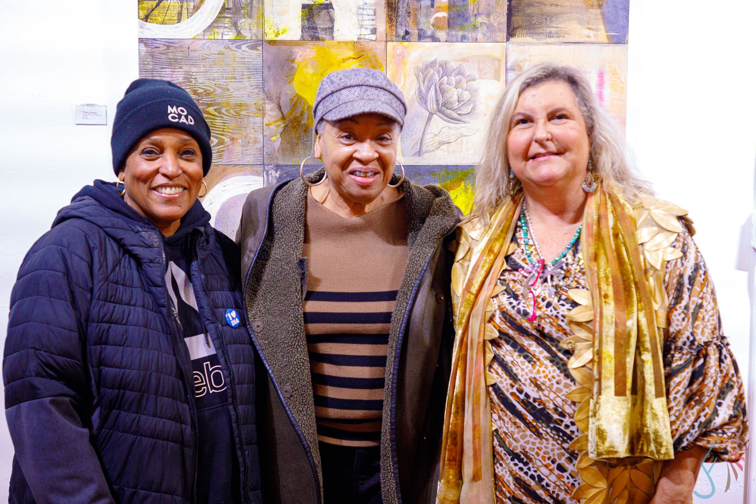 Three women smiling together at an art gallery event, wearing casual winter clothing in front of colorful artwork.