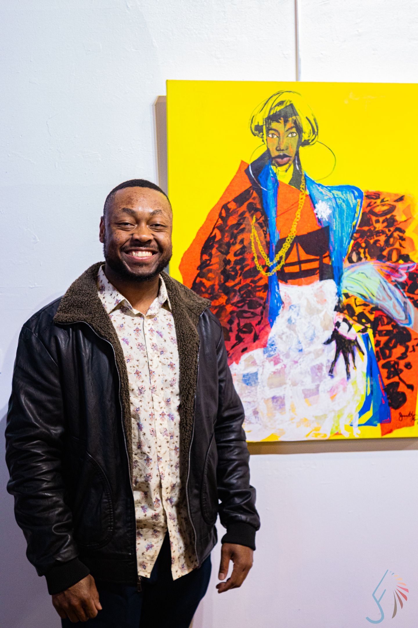 Smiling man in leather jacket stands beside vibrant portrait painting at art show.