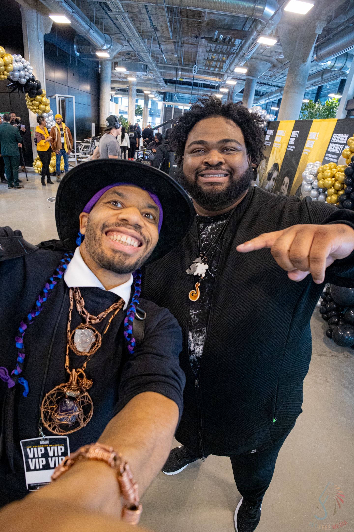 Two smiling men take a selfie at a VIP event, one wearing a purple hat and gold chains.