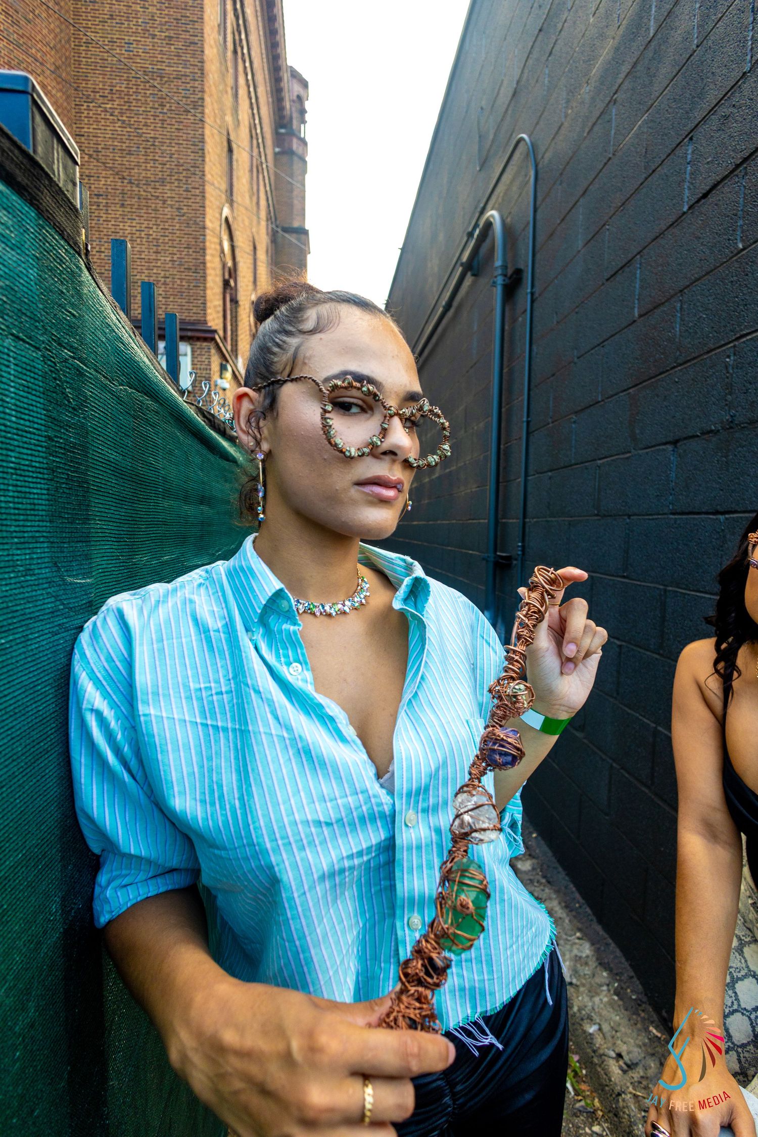 Woman in blue striped shirt with decorative sunglasses holds colorful beaded necklace in urban alley.