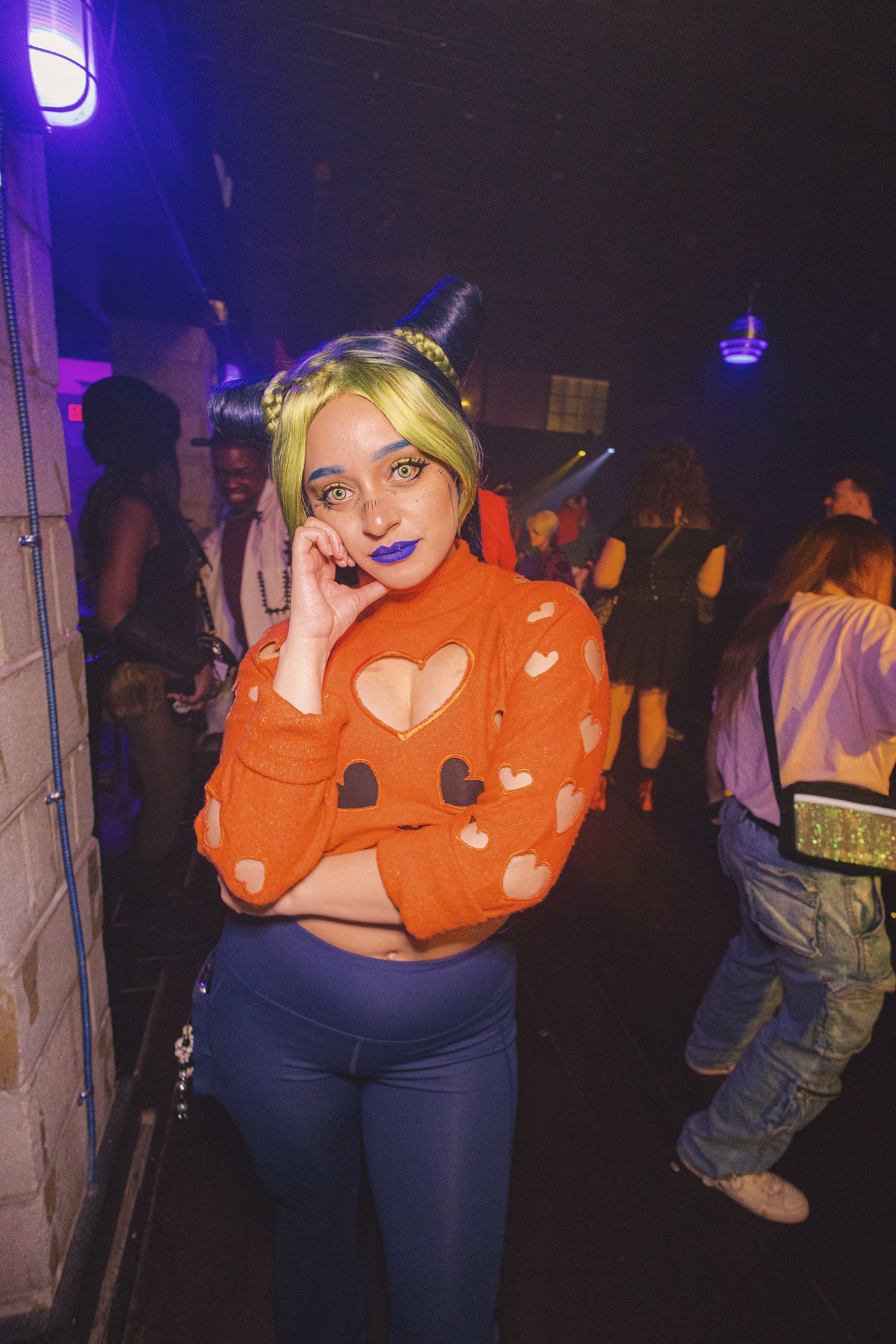 Woman in orange heart sweater with green hair and blue lipstick poses at a neon-lit party.