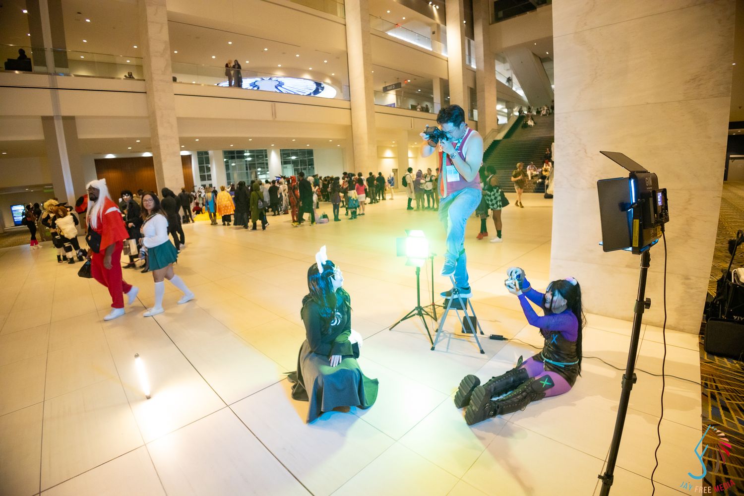 Cosplayers pose for photos in a bright convention hall with colorful costumes and studio lighting setup.