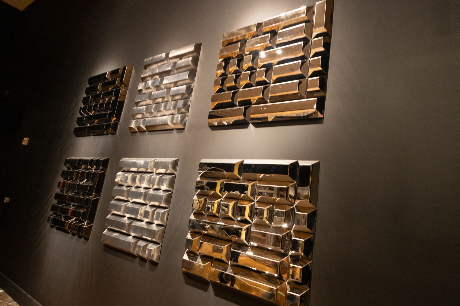 Six metallic abstract wall sculptures with geometric rectangular patterns in gold and silver tones displayed on a dark gallery wall.