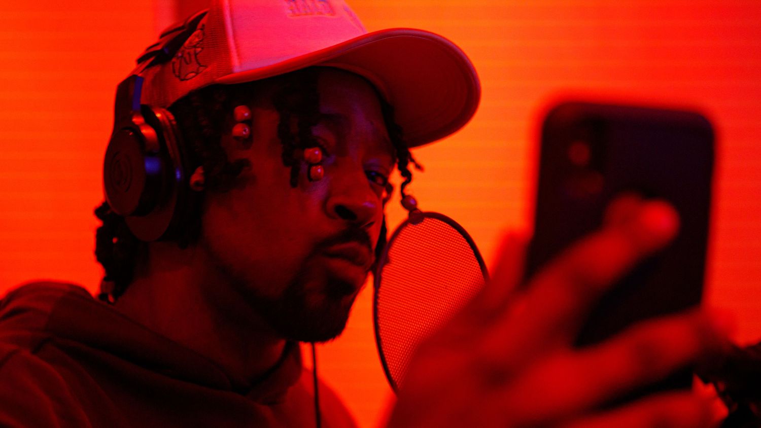 Man wearing headphones and pink cap recording music on smartphone in red-lit studio environment.