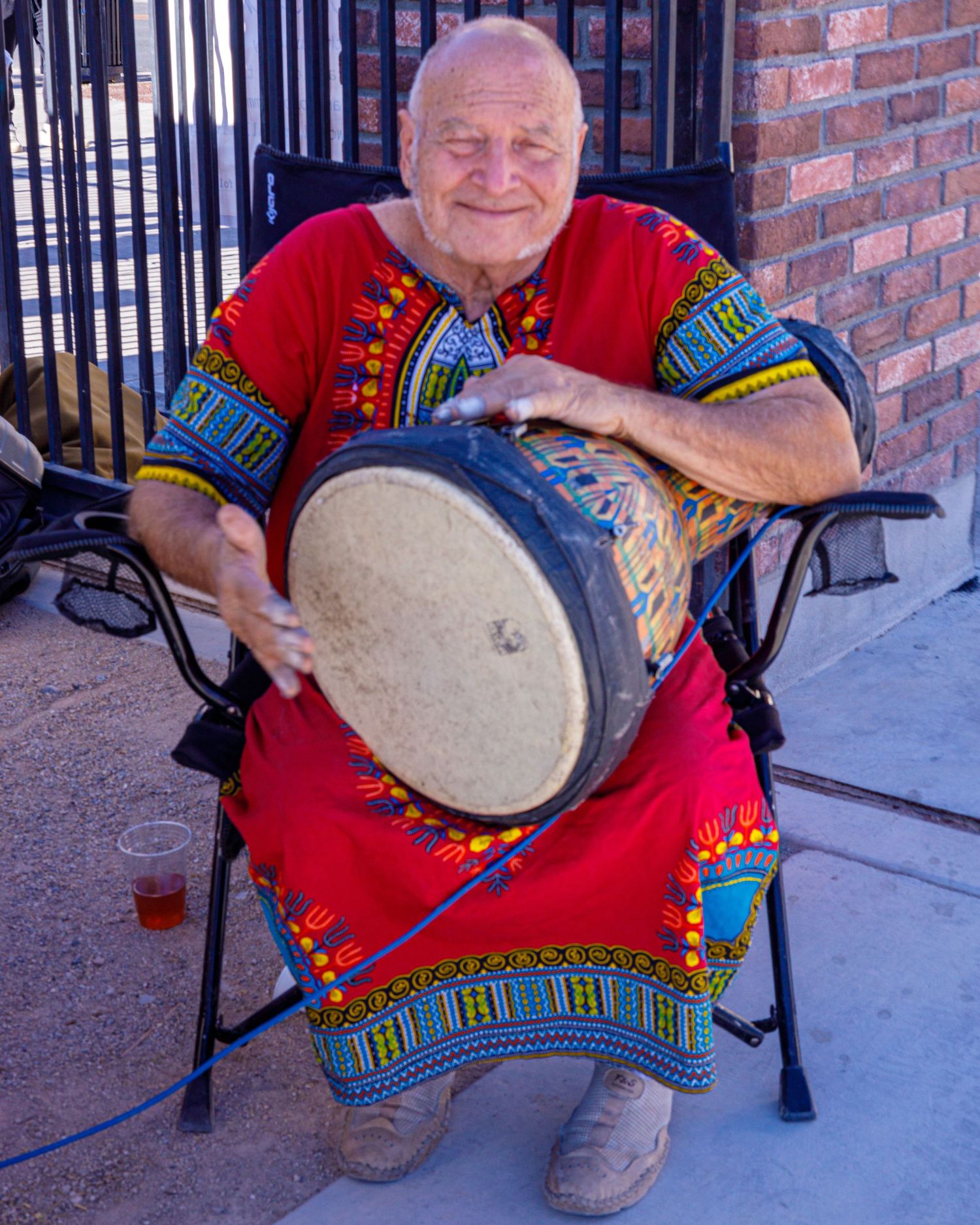 Elderly man in red dashiki plays djembe drum while seated outdoors at cultural event.
