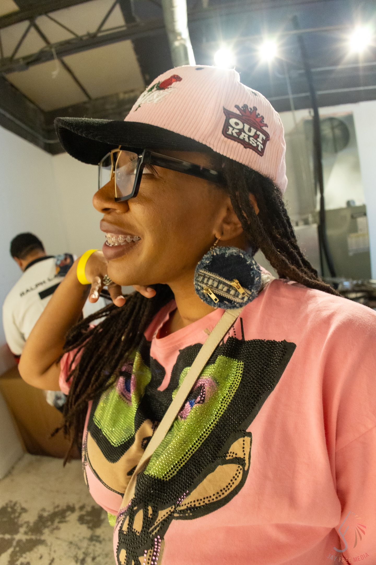 Woman smiling in pink OutKast cap, glasses, braids, and colorful earrings at an indoor event.