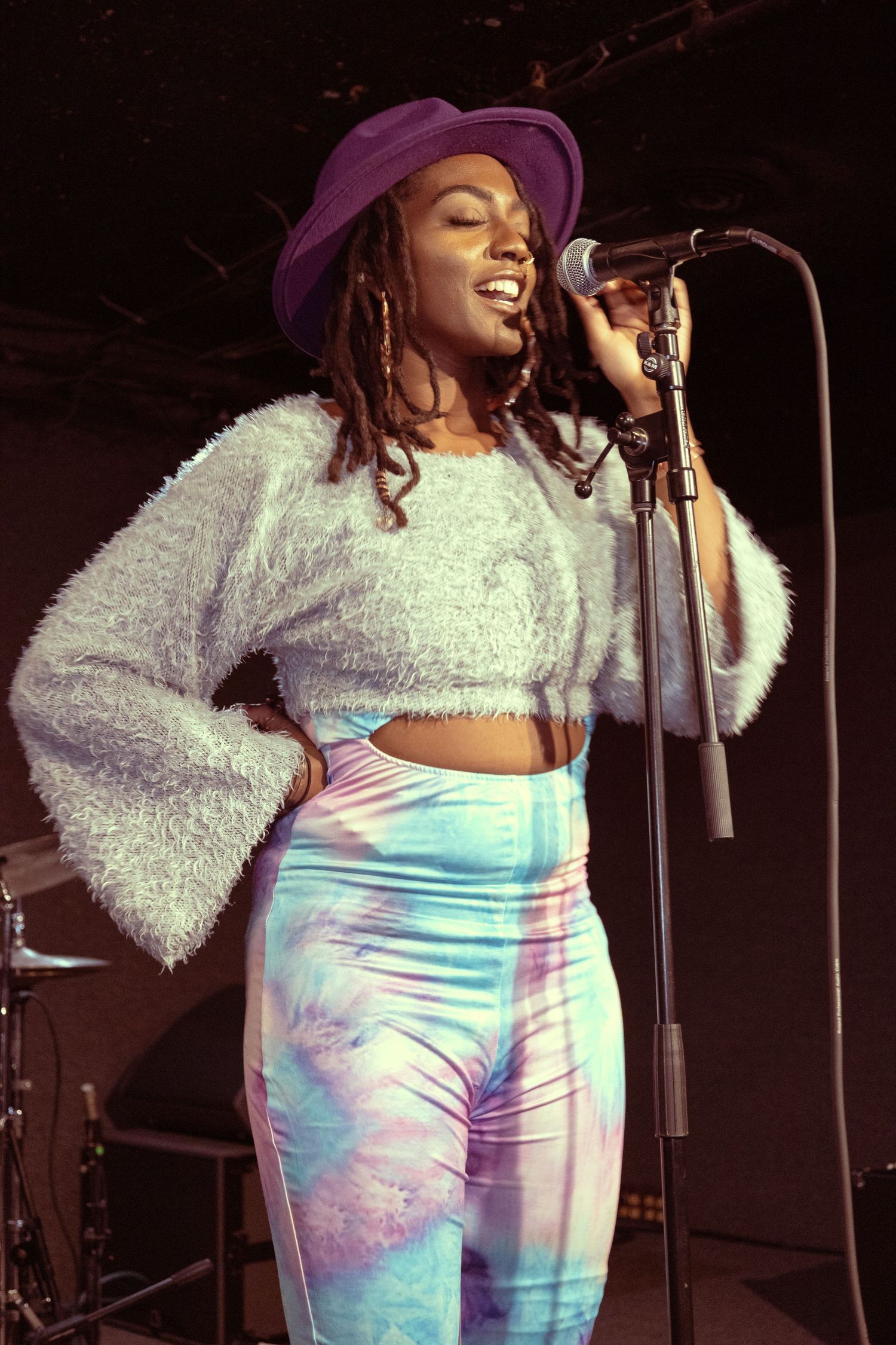 A young Black woman singer performs on stage wearing a purple hat, fuzzy crop top, and colorful tie-dye pants at a microphone.