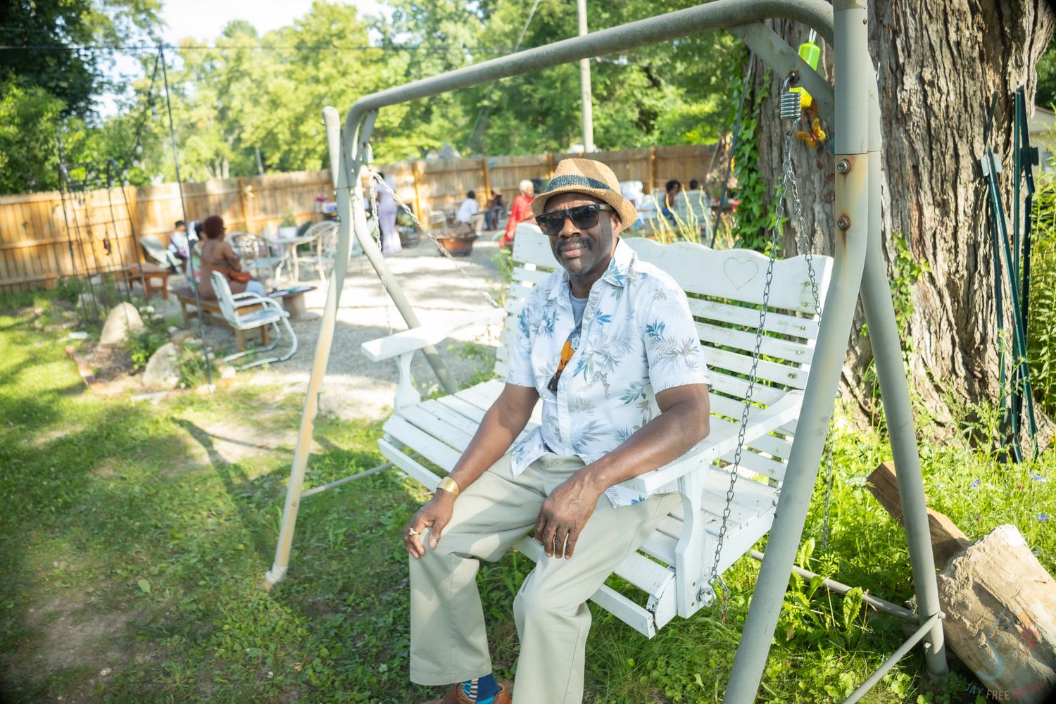A smiling man in sunglasses and a hat relaxes on a white garden swing outdoors on a sunny day.