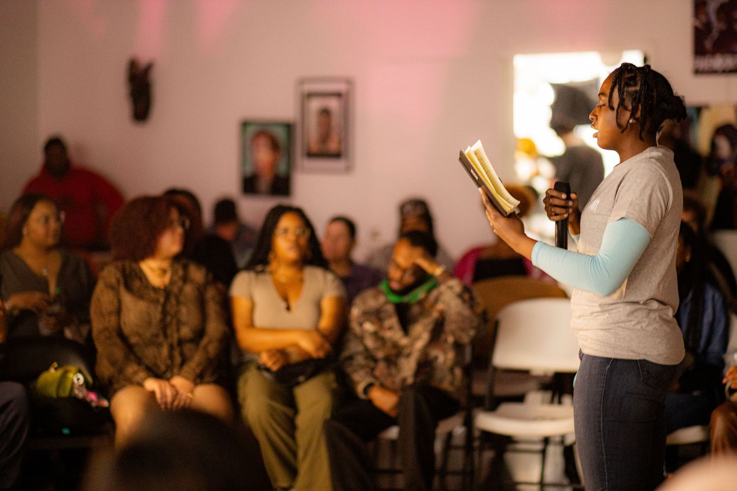 A woman reads from a book while presenting to a seated audience in a warmly lit community gathering space.