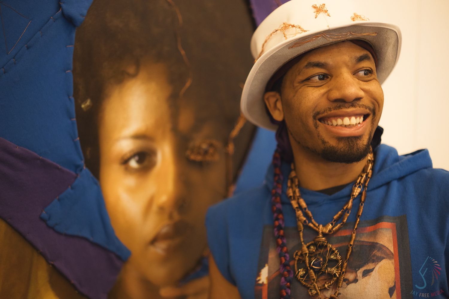 A person in a blue hoodie and white hat smiles while standing next to a colorful wall mural.