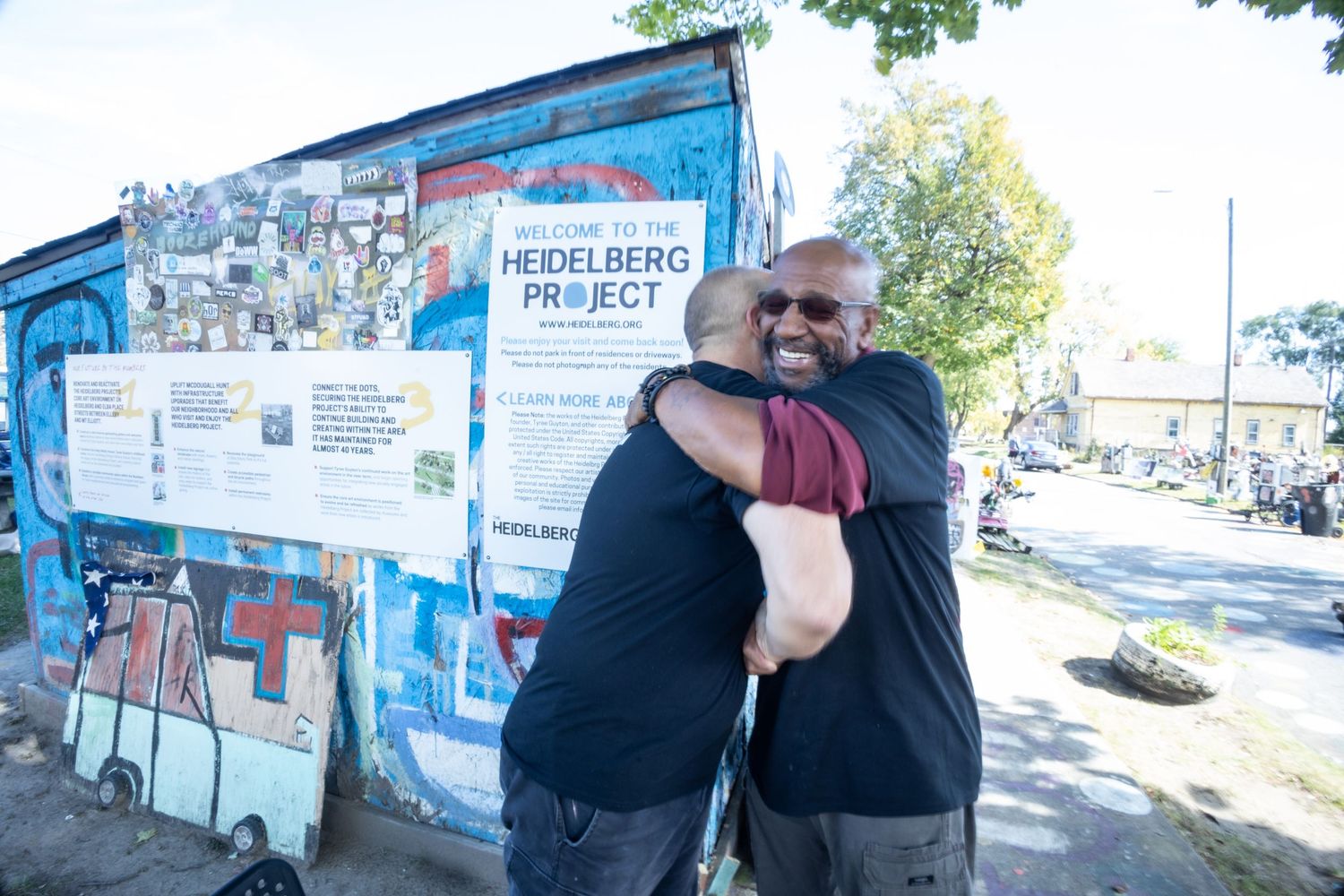 Two men hugging warmly in front of the Heidelberg Project outdoor art installation in Detroit, Michigan.