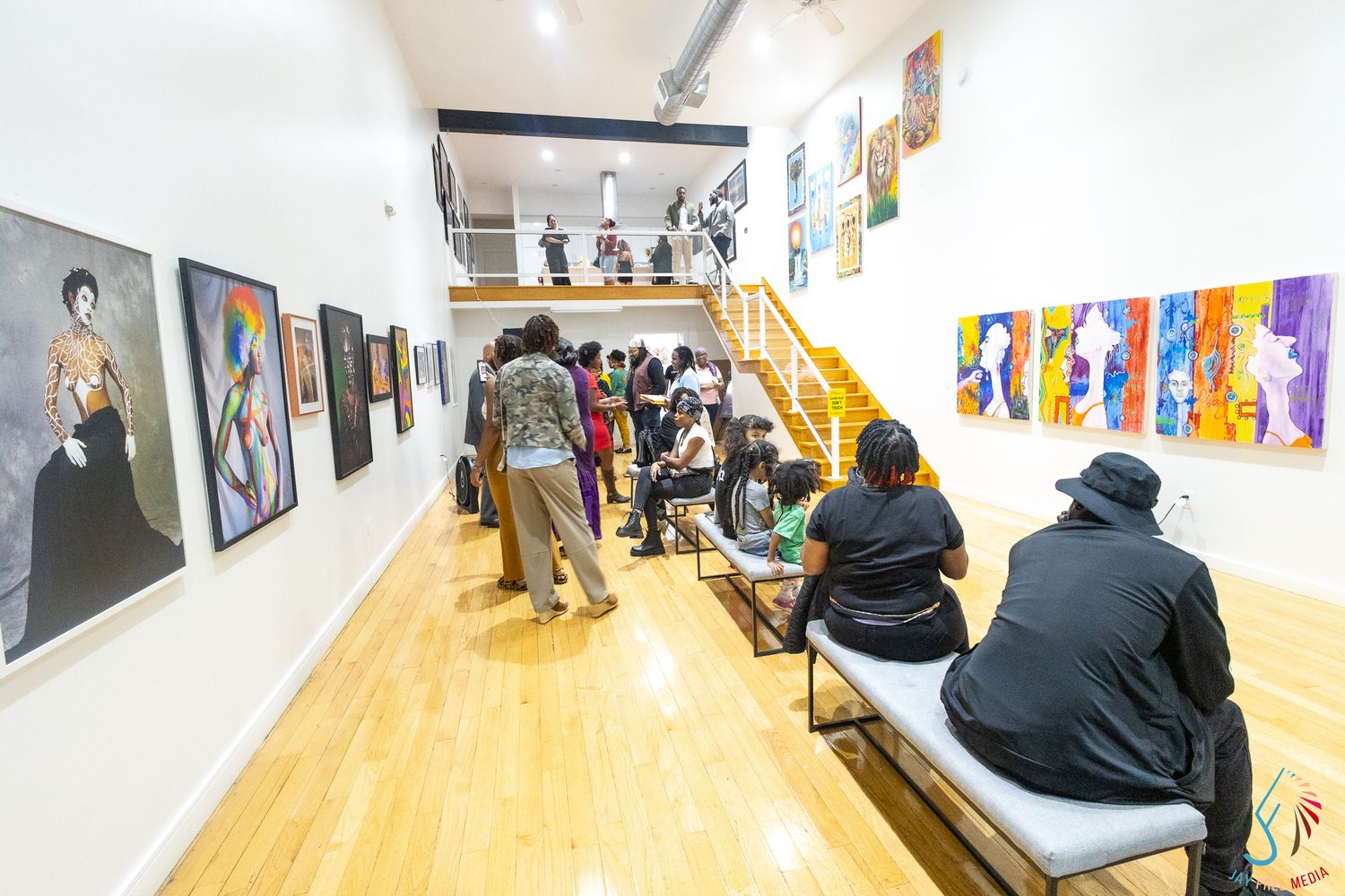 Visitors explore a bright art gallery with colorful paintings on white walls and a staircase in the background.