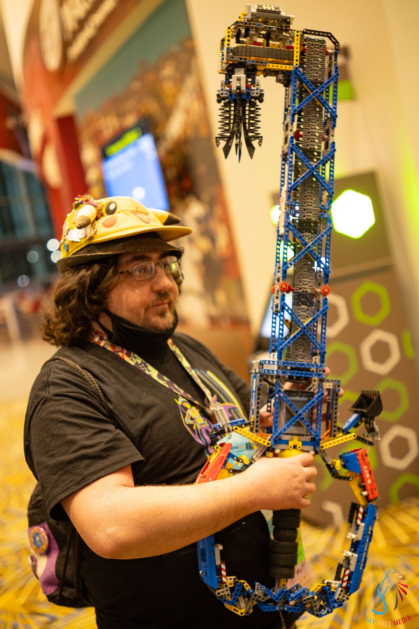 Man holding large K'Nex guitar sculpture wearing decorated hard hat at gaming convention.