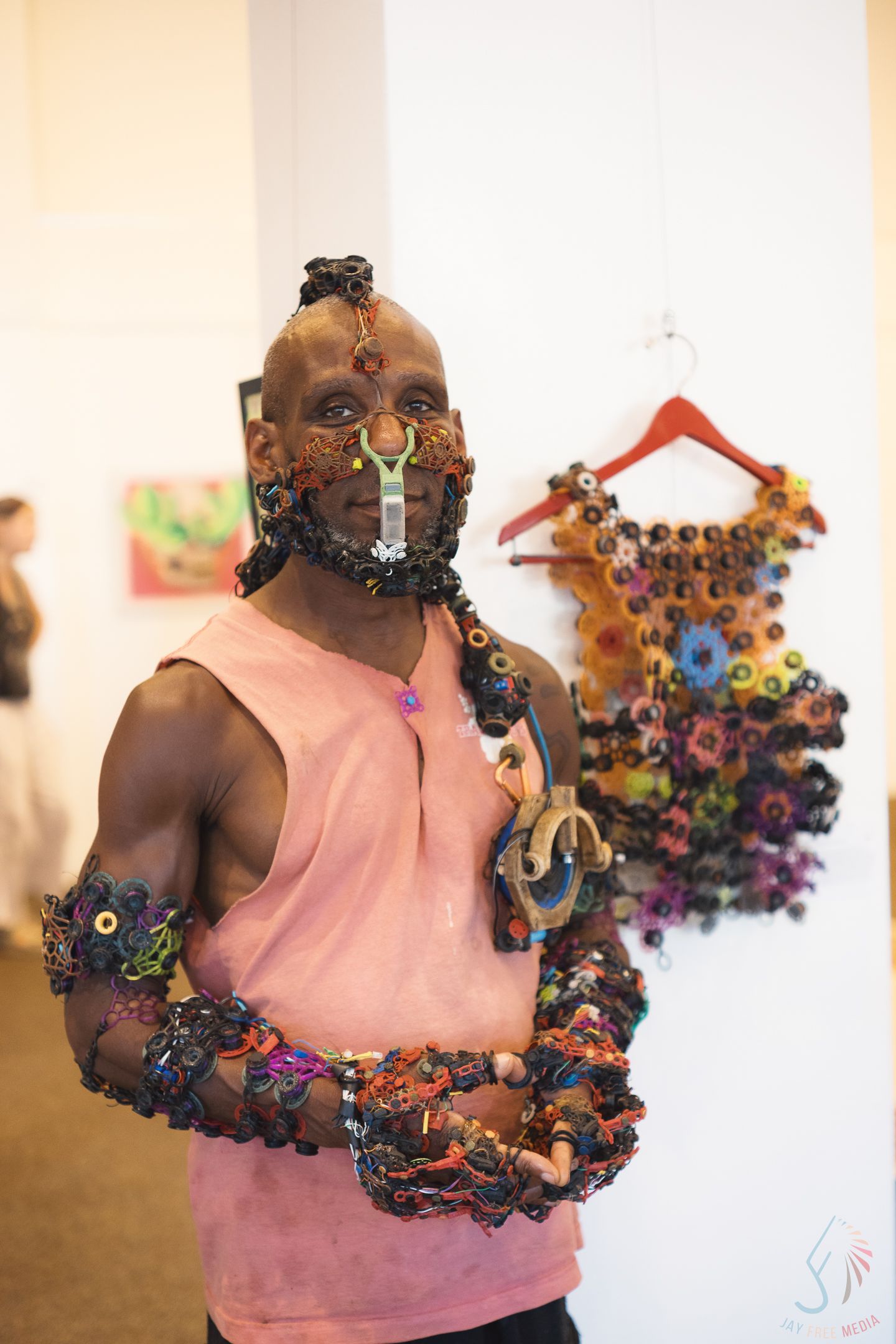 A man wearing elaborate colorful beaded accessories on his face and arms stands in an art gallery.