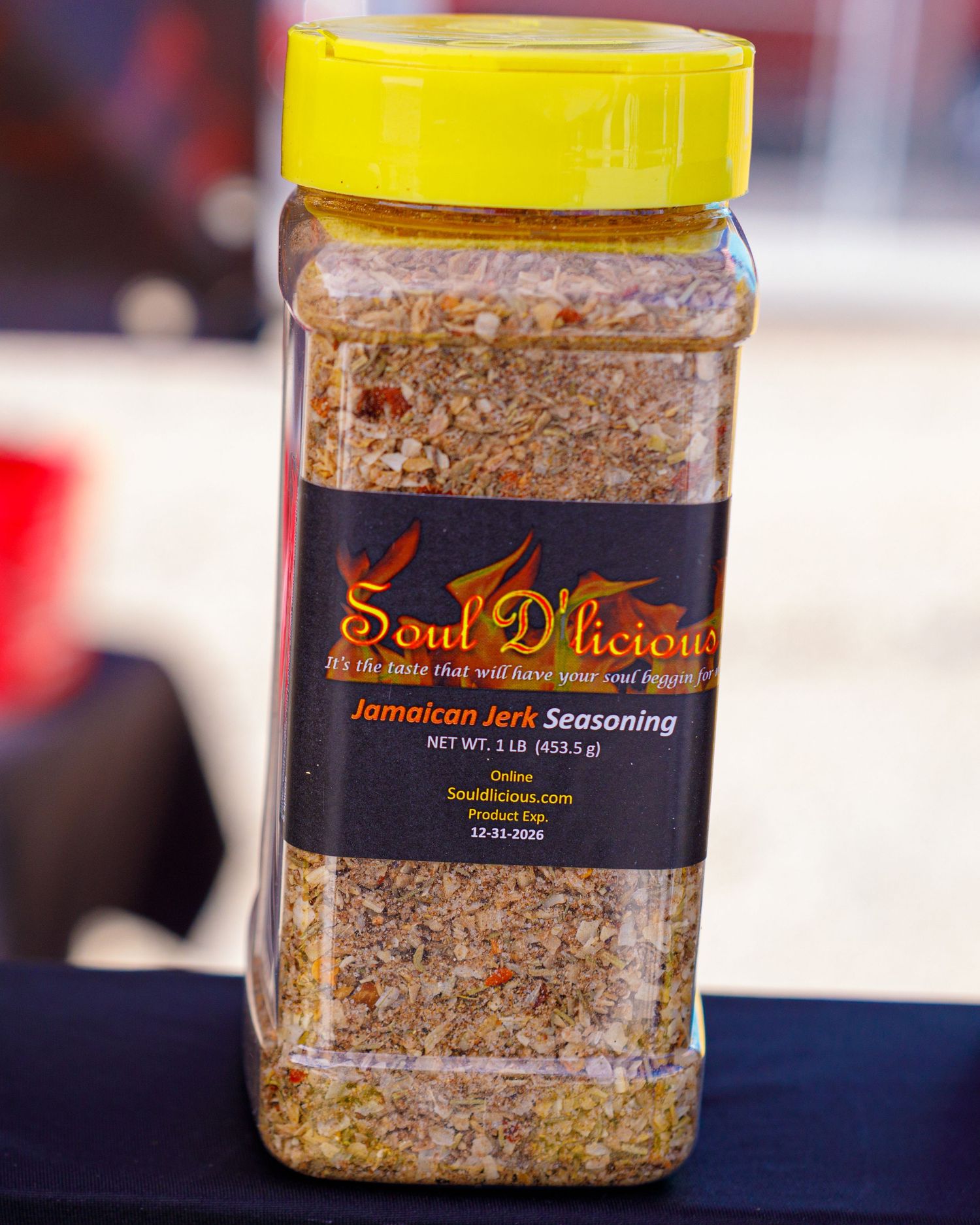 A large jar of Soul D'licious Jamaican Jerk Seasoning with a yellow lid at an outdoor market.