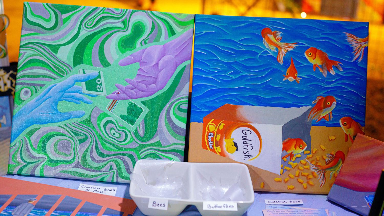 Two colorful paintings displayed at a market, featuring abstract swirls and vibrant goldfish art.