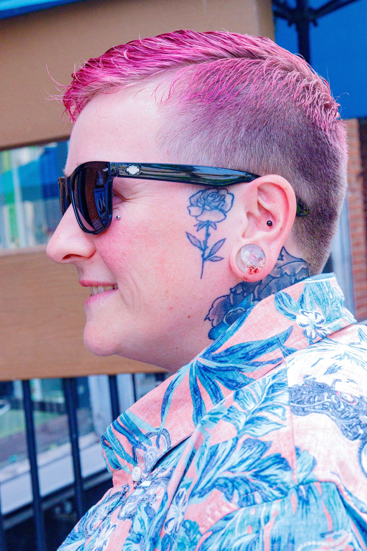Person with pink undercut hair, sunglasses, and floral neck tattoo wearing a tropical shirt outdoors.