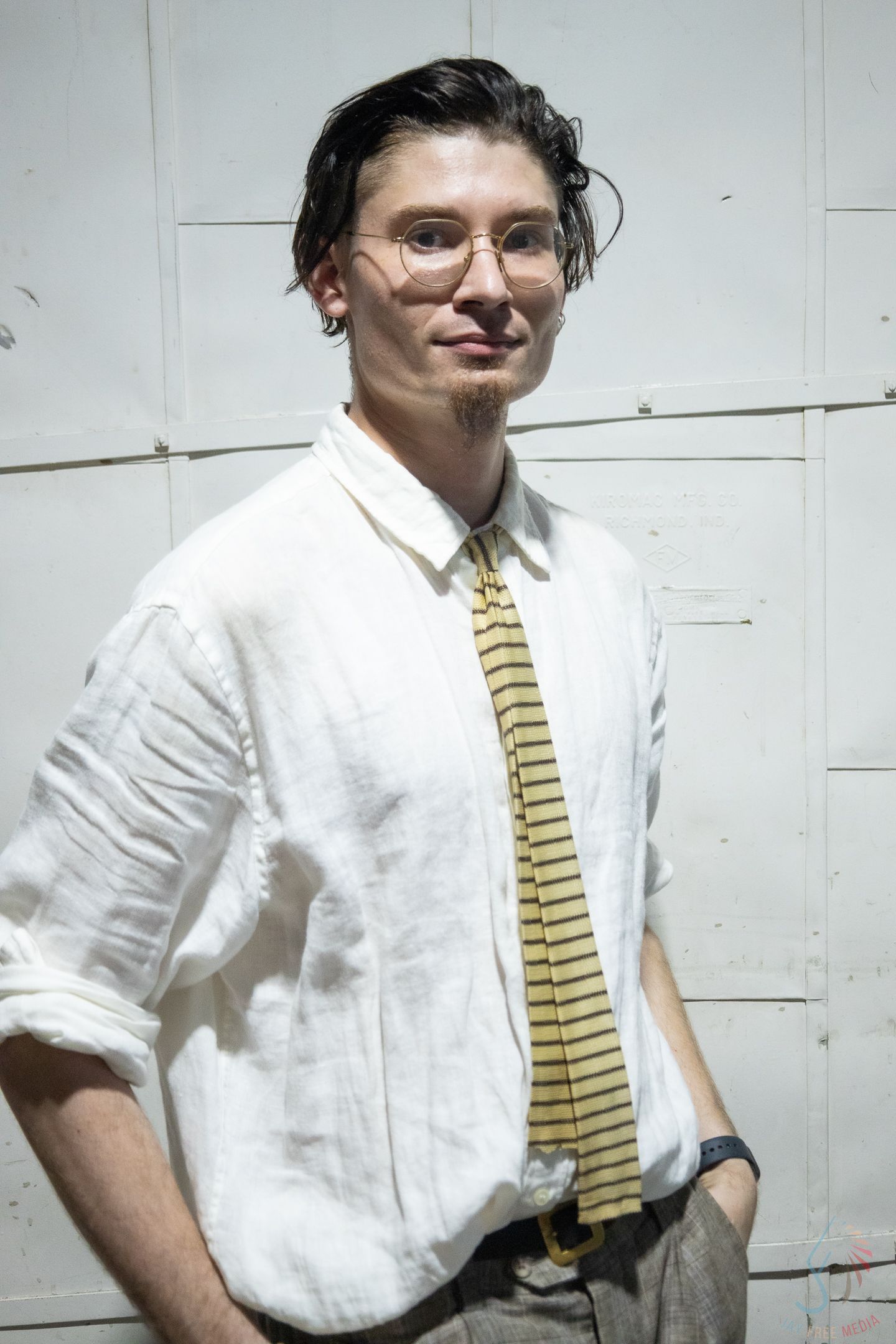 Young man with glasses wearing a white linen shirt and striped yellow tie, smiling against a white wall.