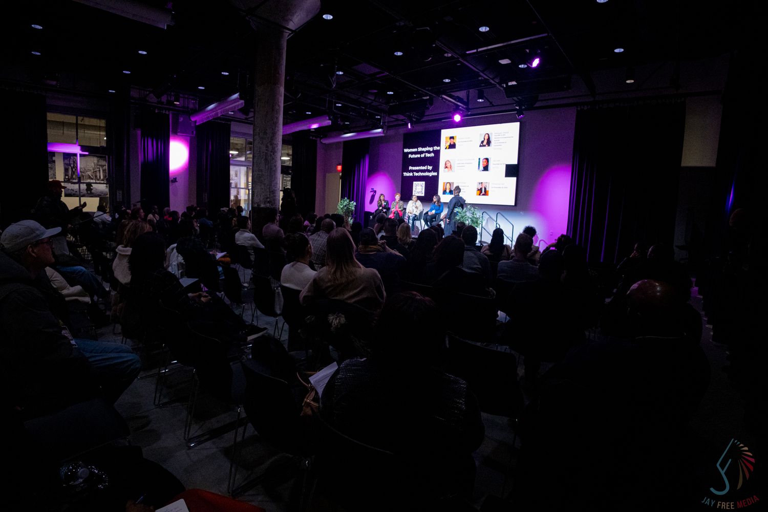 Audience watches panel discussion on stage with purple lighting at an indoor conference event.