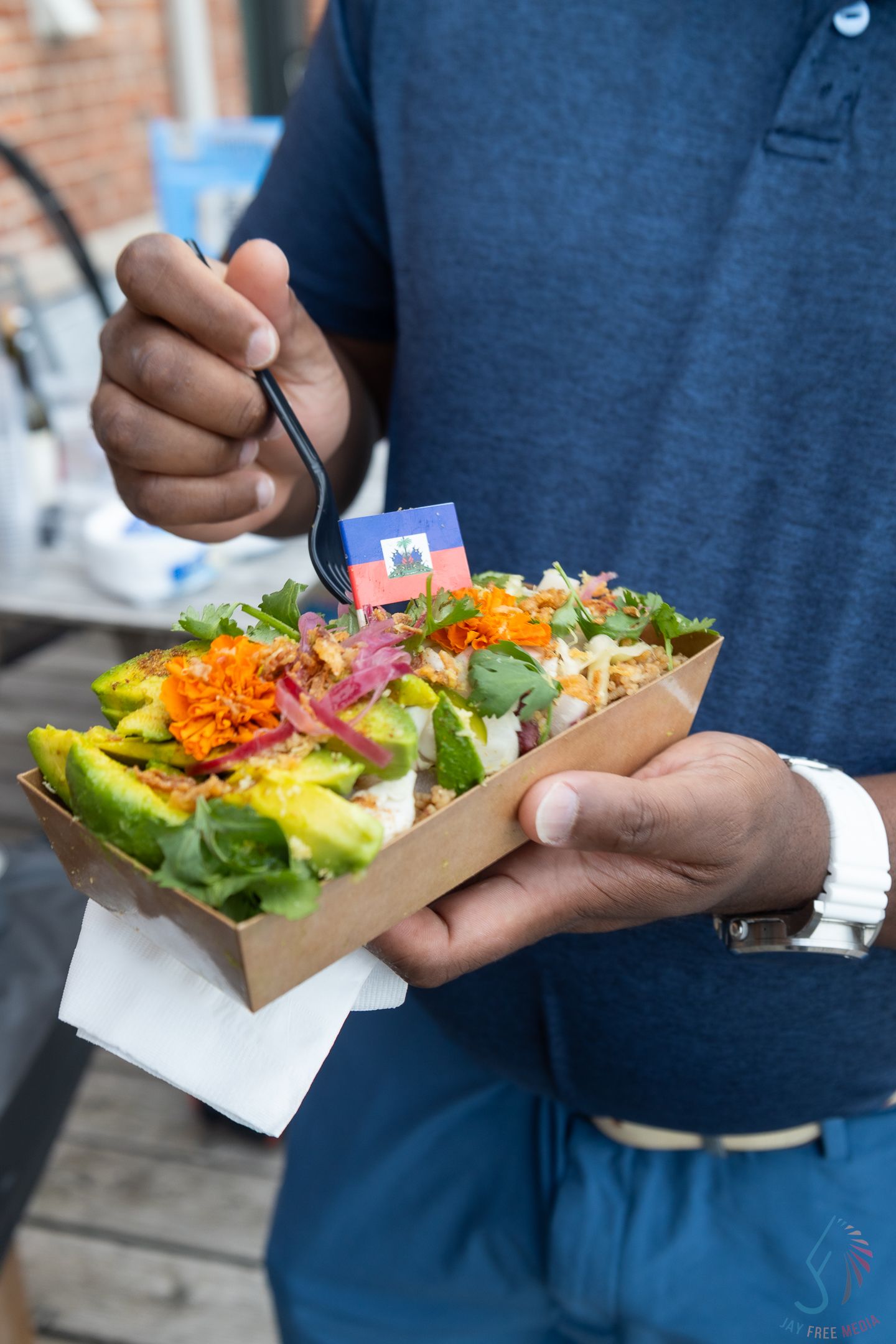 Man in blue shirt holding a fresh salad bowl with avocado, carrots, and greens, eating with a fork outdoors.
