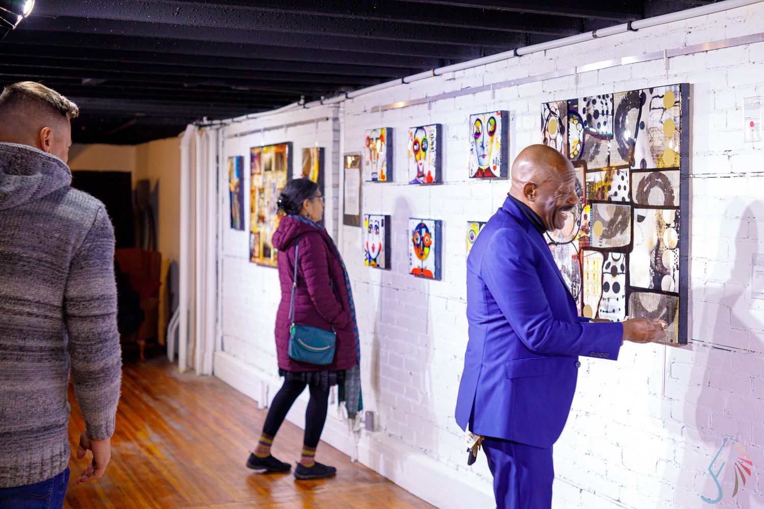 People viewing colorful artwork displayed on white brick walls in a modern art gallery with hardwood floors.