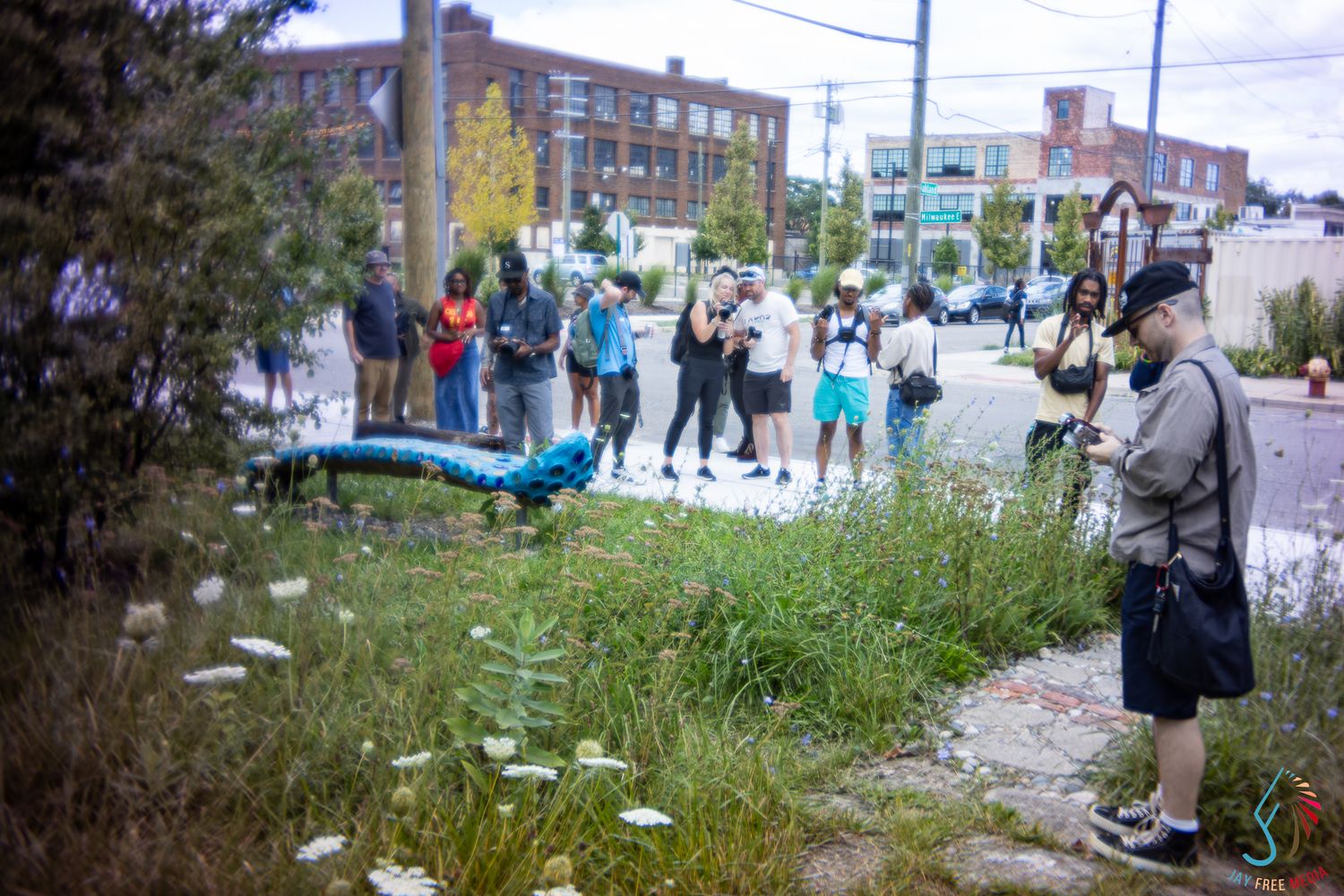 Group of people gathered outdoors in an urban lot viewing a blue art sculpture in tall grass.