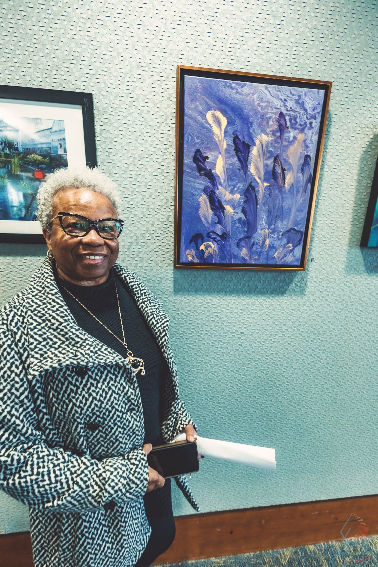 Smiling woman with white hair and glasses stands beside blue abstract painting at an art gallery exhibition.