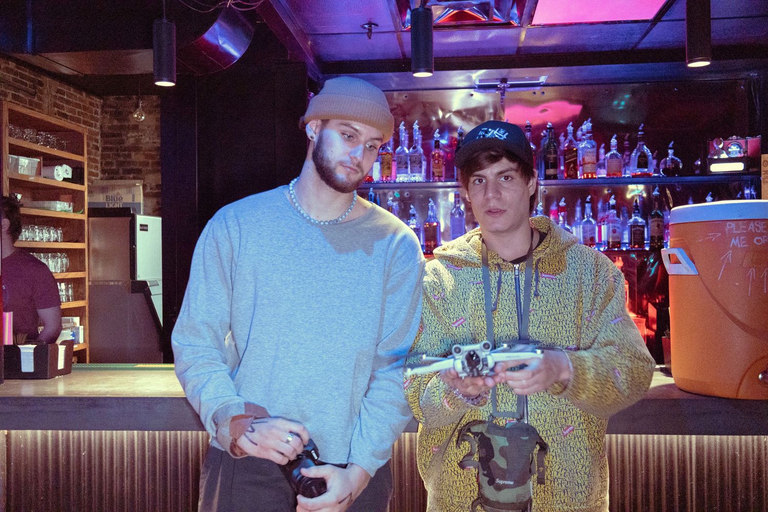 Two young men standing in a dimly lit bar, one holding a camera and the other a drone controller.