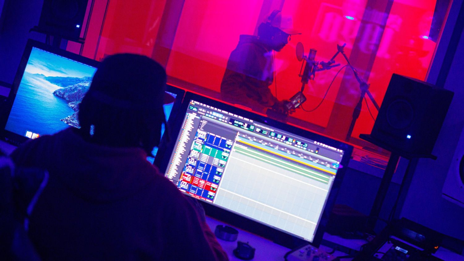 Music producer at a computer workstation records an artist singing in a studio bathed in red and blue lighting.