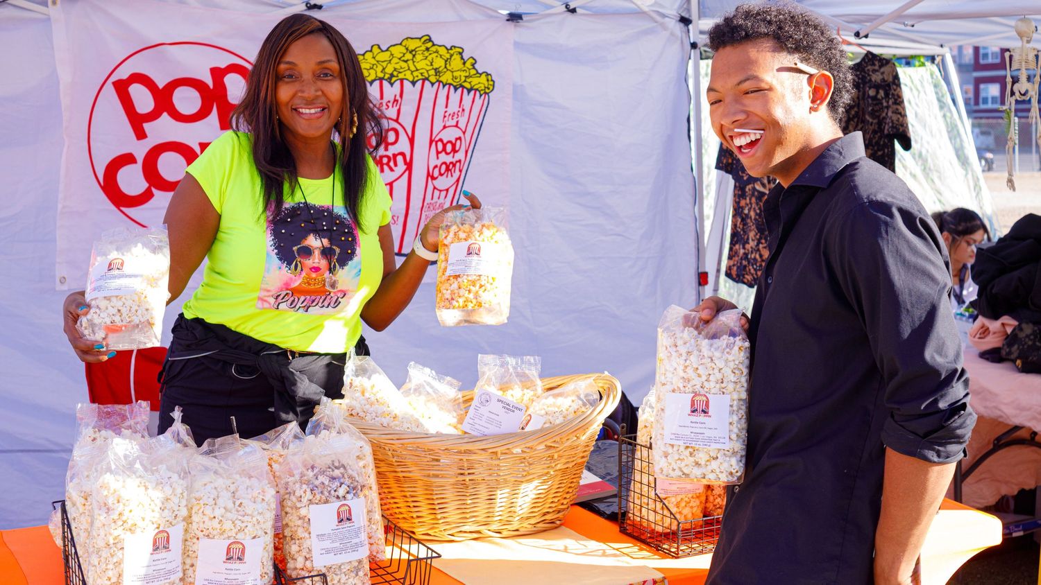 Two vendors smiling at popcorn stand with bags of gourmet popcorn at outdoor market.