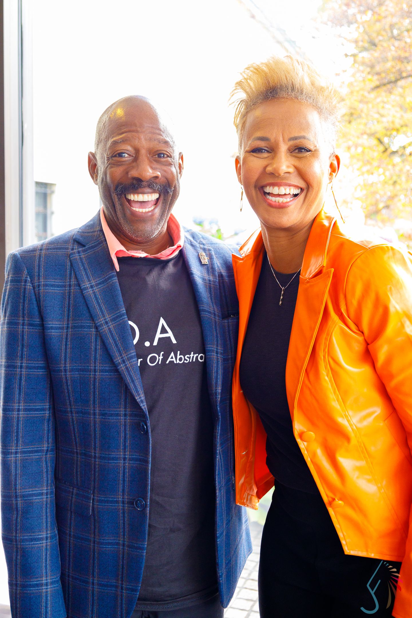 Smiling man in blue plaid blazer and woman in orange leather jacket pose together at sunny event.