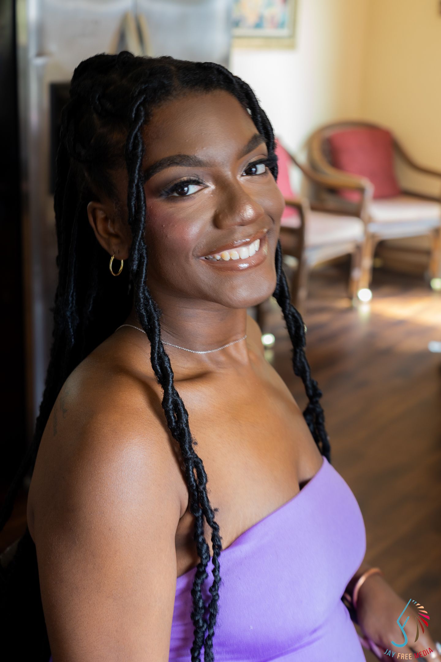 Smiling young Black woman with braided hair wearing a purple dress and hoop earrings at an indoor event.